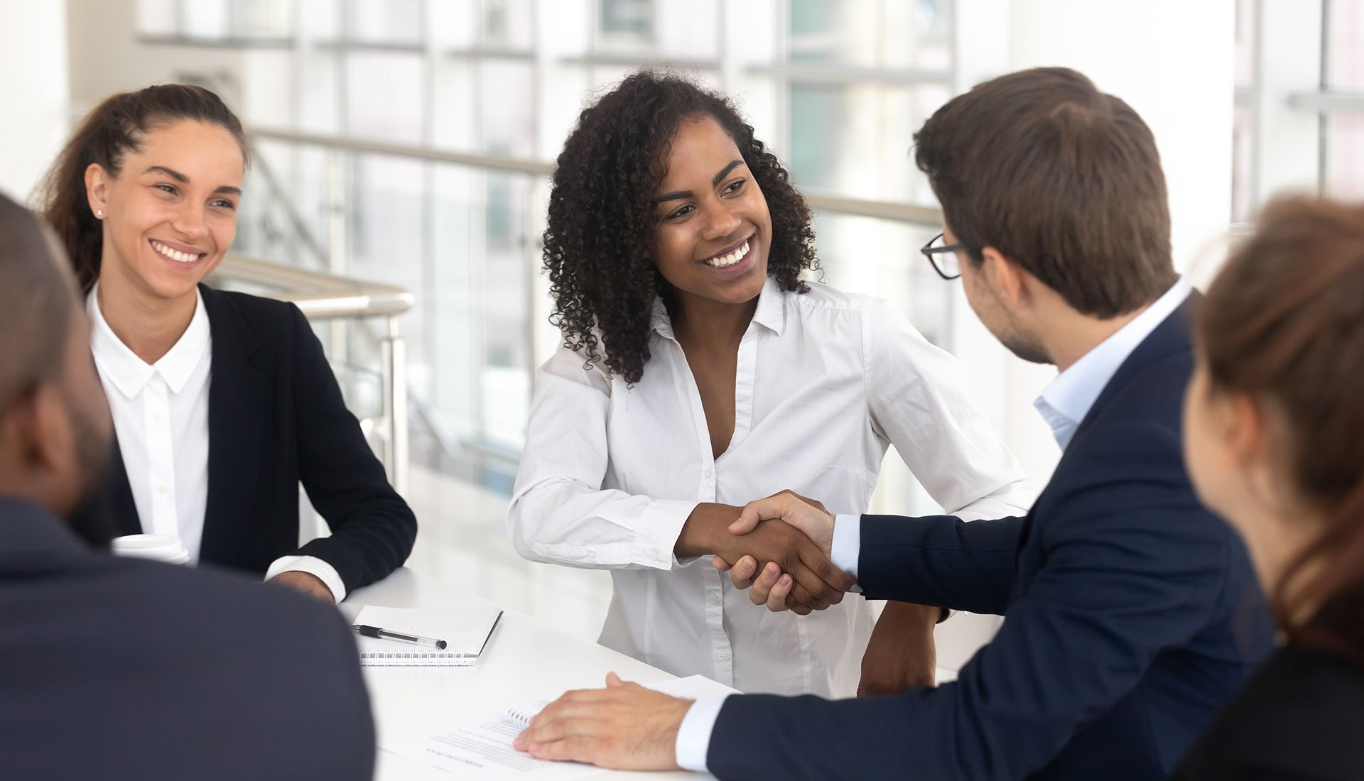 Businesspeople shake hands at a table in an office, smiling.