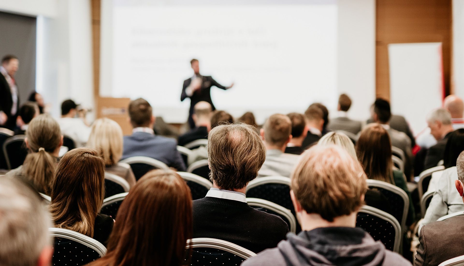 Audience seated, listening to a presentation in a brightly lit conference room. Speaker is standing by a screen.