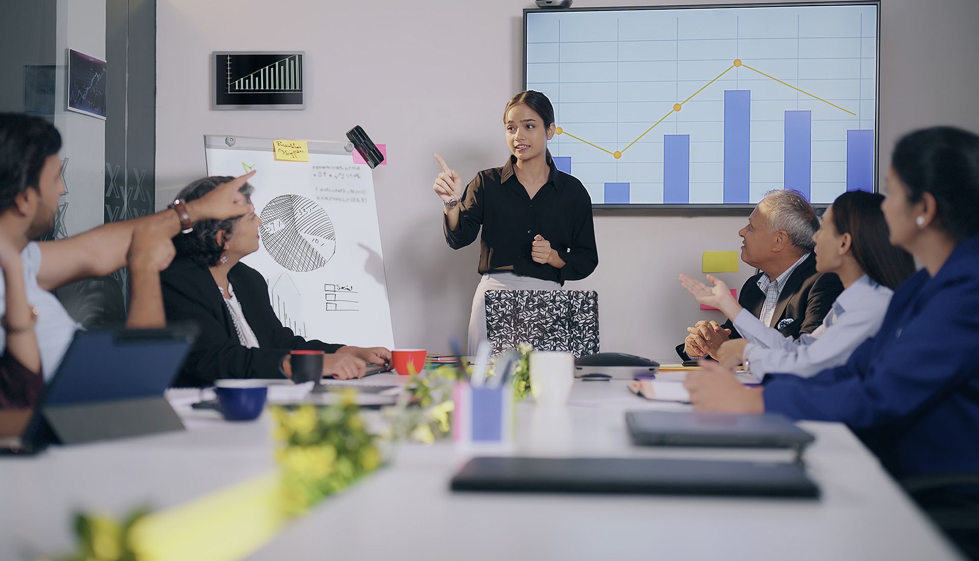A woman leads a meeting in a conference room. Attendees sit around a table, and a chart is displayed.