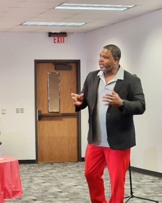 Man speaking in a room, gesturing beside a wooden door and red tablecloth.