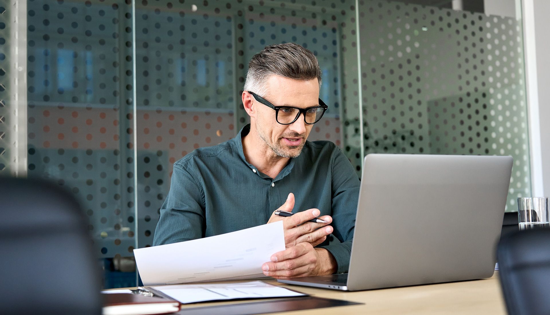 Man in glasses looking at papers and laptop in an office.