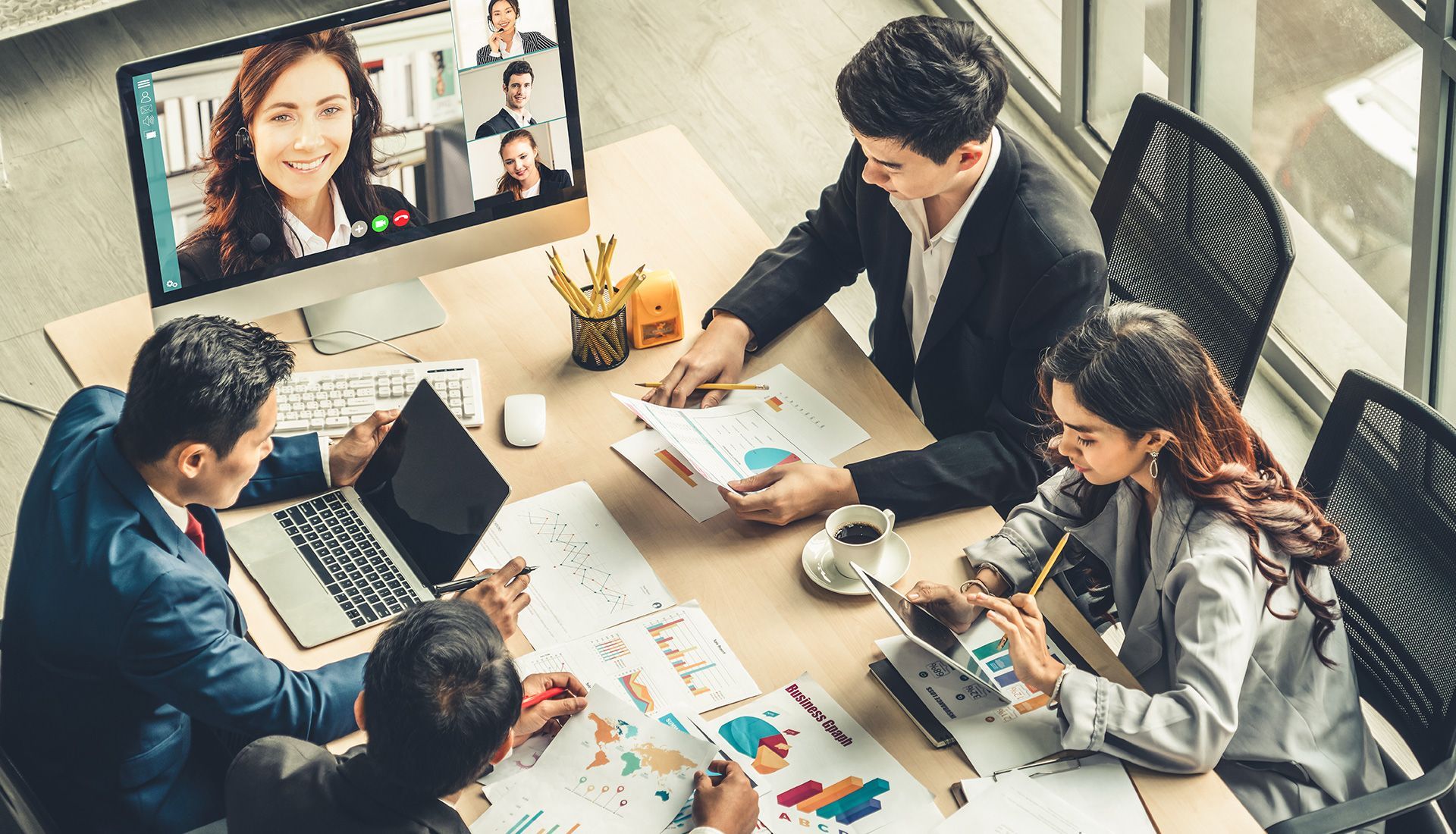 Business team in a meeting with remote participants on a computer screen, reviewing documents and using technology.