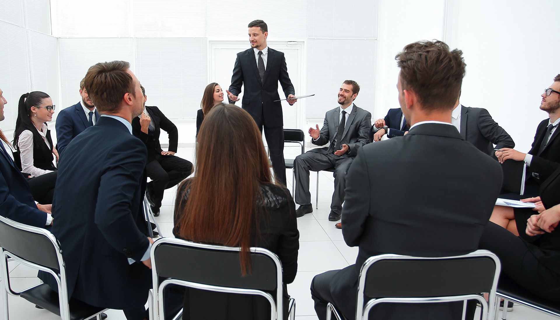 Group of people in suits in a meeting; one man stands and speaks while others sit, listening.