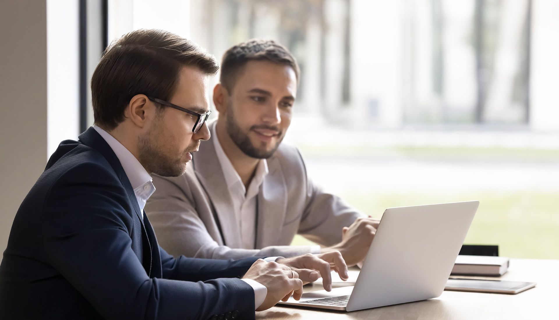 Two men in suits, focused on a laptop screen in a bright office, one typing and the other pointing.