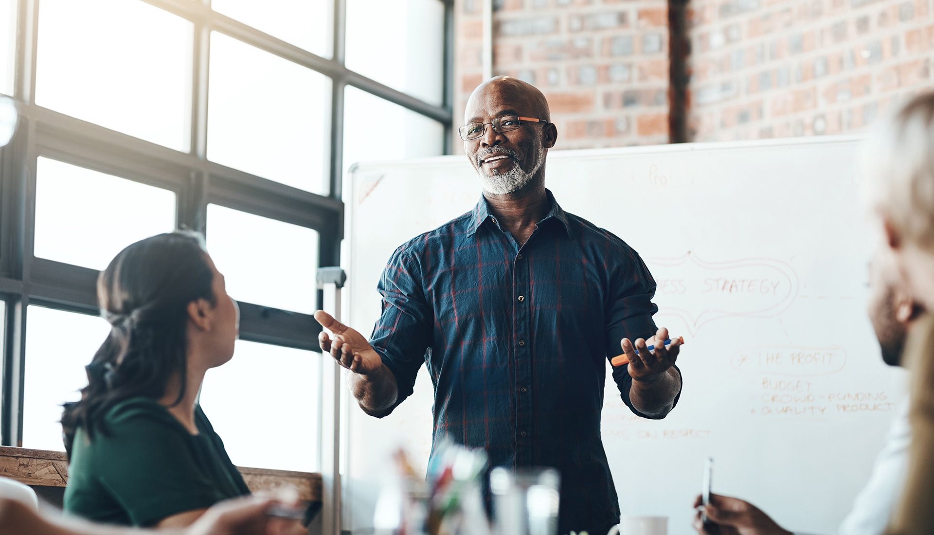 Man speaking in meeting, gesturing. Whiteboard, office setting.
