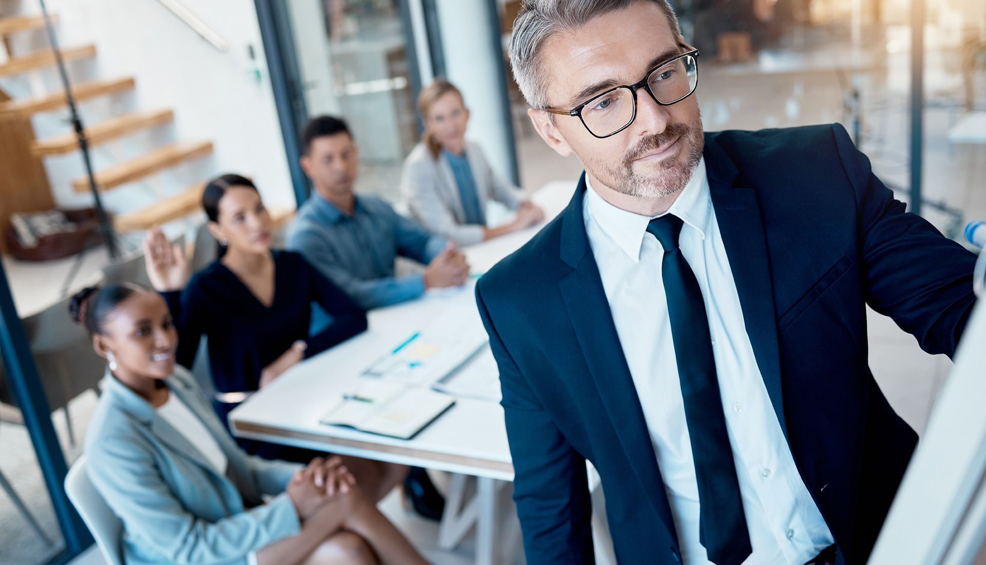 Man in suit leading a presentation in an office; colleagues listening.
