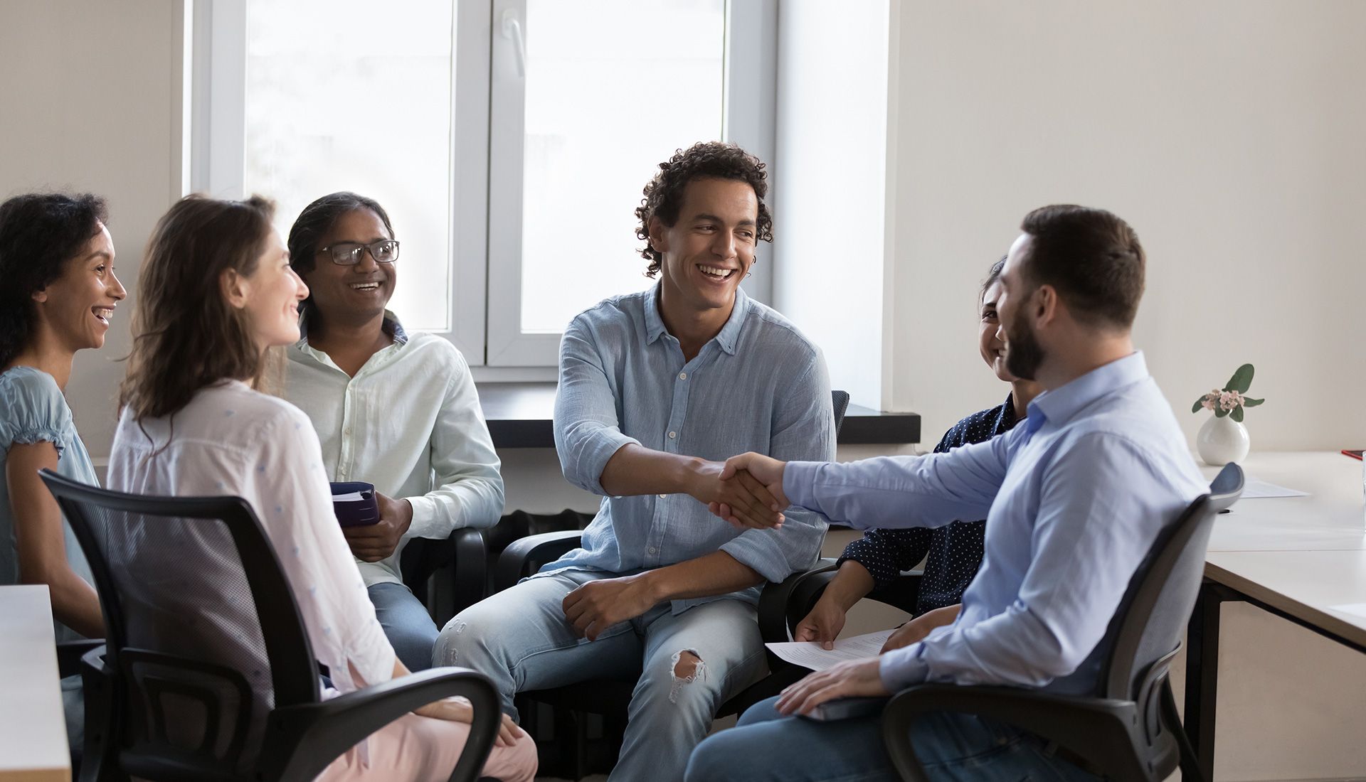 Group of people in an office. Two men shake hands; others look on, smiling.