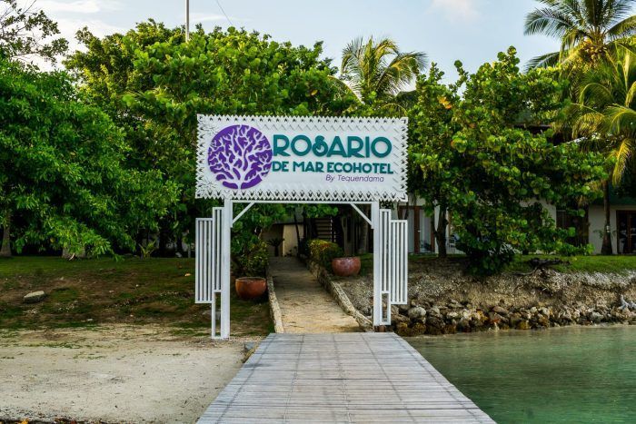 Letrero para el Ecohotel Rosario de Mar, entrada por un muelle de madera, con exuberante vegetación verde.
