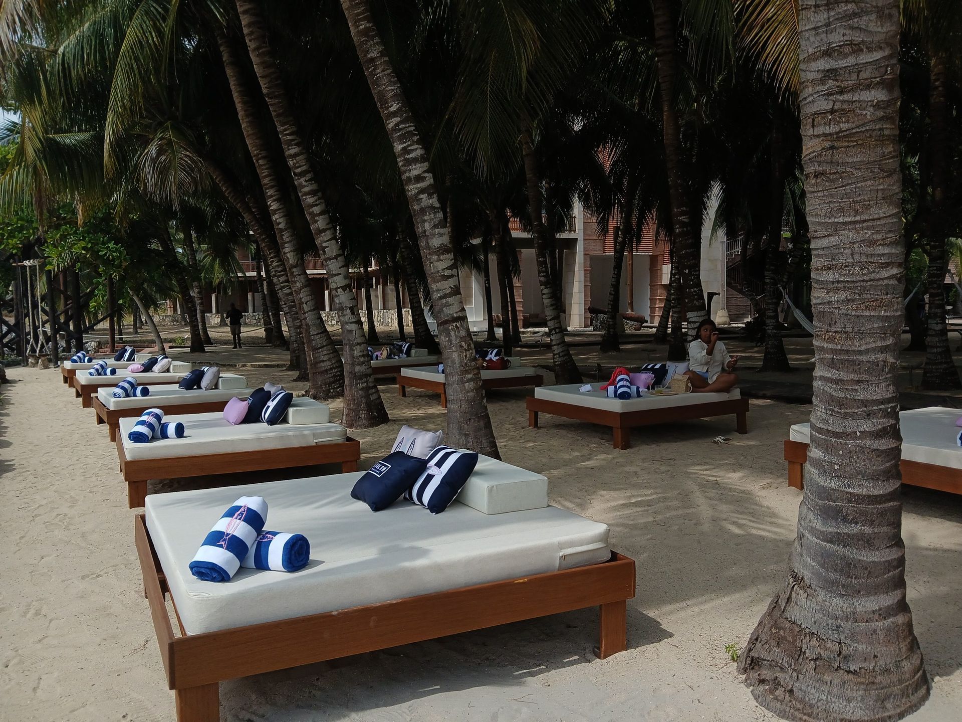 Tumbonas de playa bajo palmeras. Cojines blancos, toallas a rayas azules y suelo arenoso.