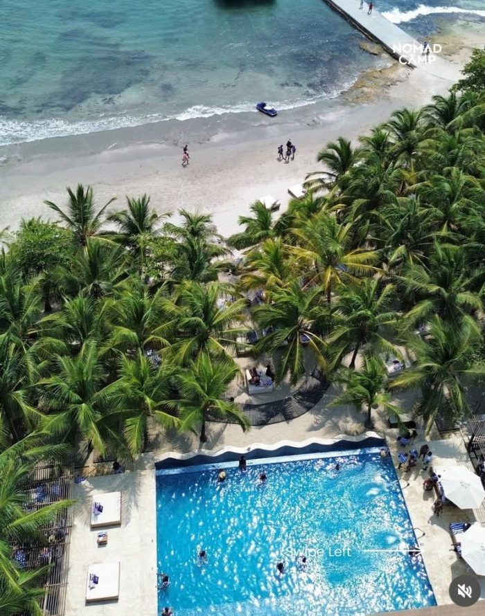 Vista aérea de la playa con palmeras, piscina y gente nadando y caminando. Agua azul y arena blanca.