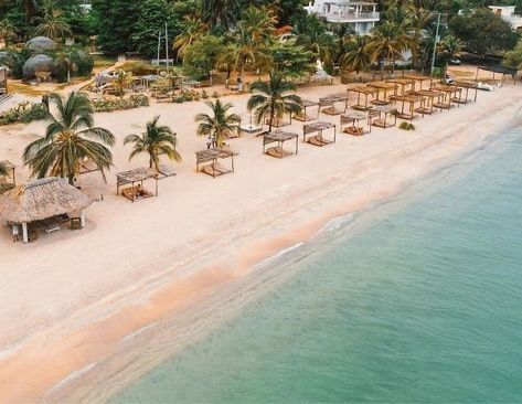 Playa con palmeras, cabañas y agua turquesa. Orilla arenosa con un edificio al fondo.