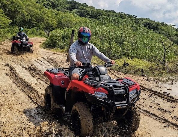 Dos personas conducen quads rojos por un sendero fangoso en una zona boscosa.
