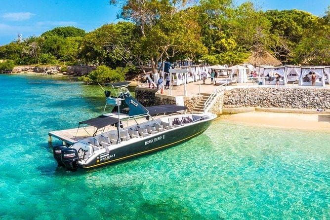 Barco en una playa tropical de aguas turquesas; gente en la orilla con cabañas blancas.