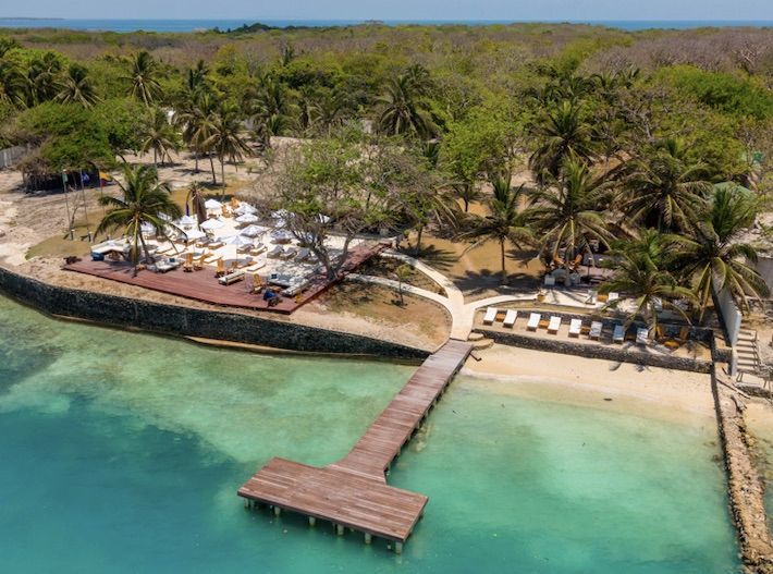 Vista aérea de un muelle de madera, playa y tumbonas con el océano y los árboles de fondo.