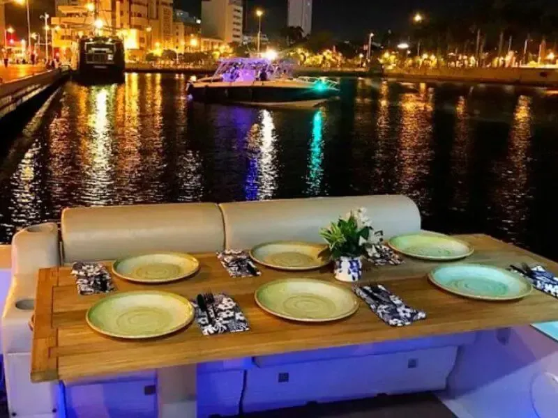 Una mesa puesta para una comida en un barco por la noche, reflejando las luces de la ciudad en el agua.