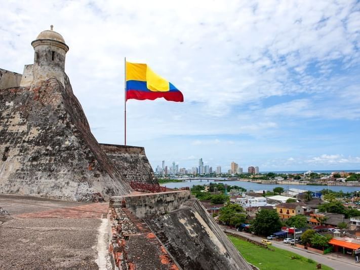 Bandera colombiana ondeando en lo alto de una fortaleza de piedra con vistas a una ciudad y a la costa.