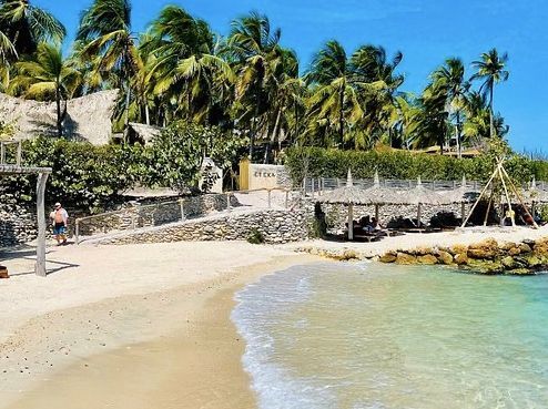 Playa de arena con agua cristalina, palmeras y pequeñas construcciones. Una persona camina por la orilla.