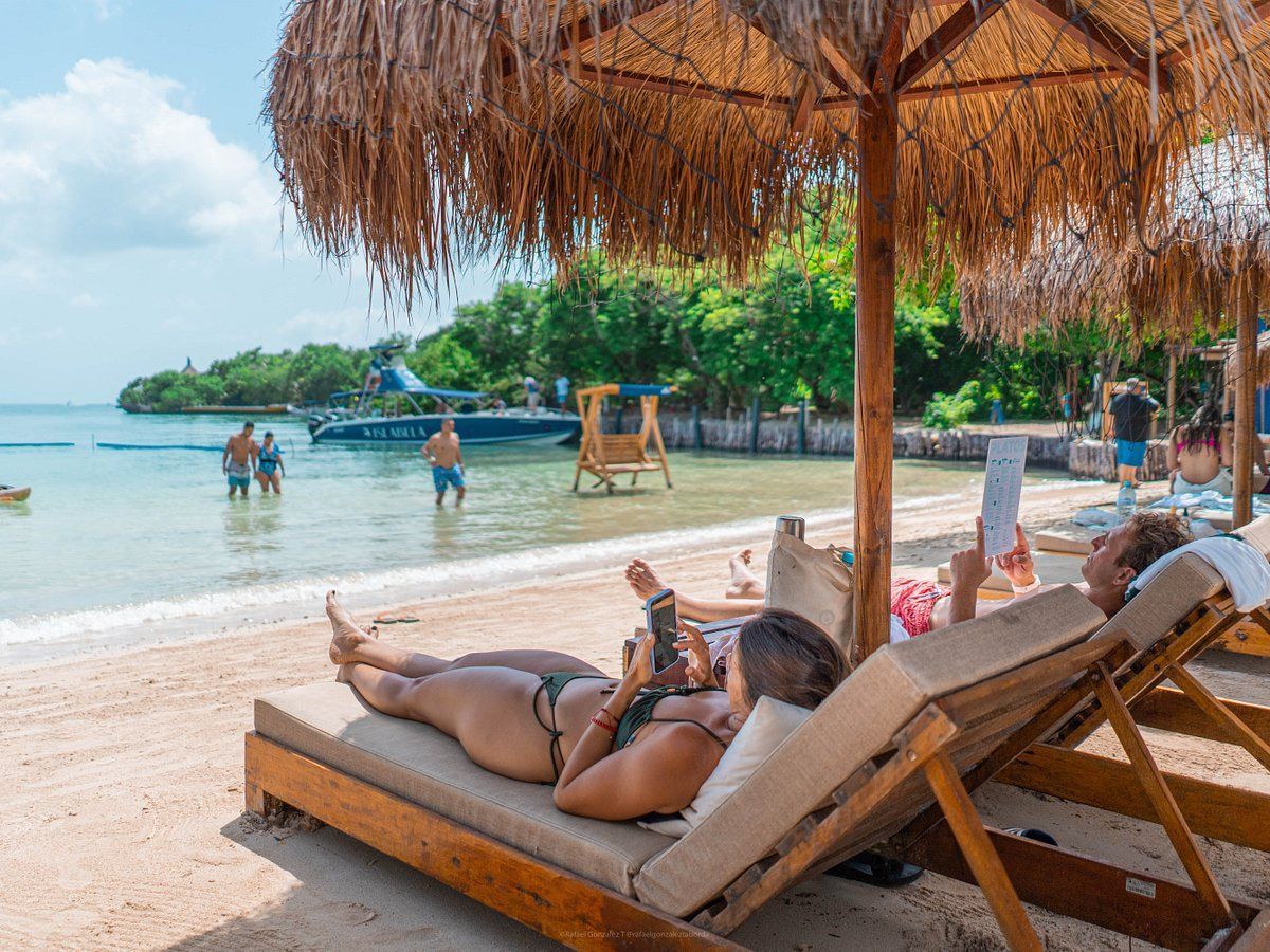 Escena de playa: Personas relajándose en tumbonas bajo sombrillas, cerca del agua. Barco y bañistas al fondo.