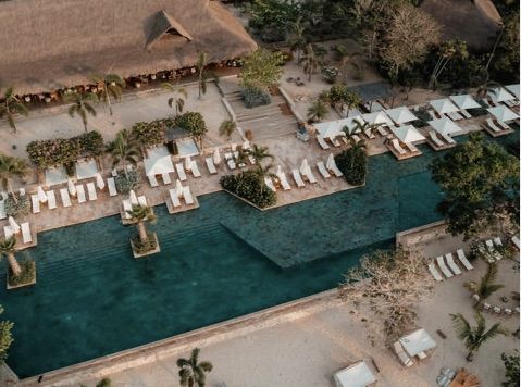 Vista aérea de un complejo turístico con una larga piscina, tumbonas blancas, techos de paja y palmeras.
