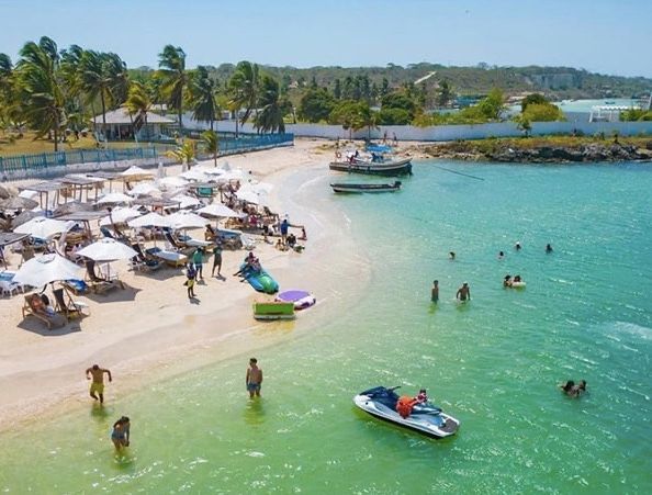 Escena de playa con gente nadando, tomando el sol bajo sombrillas y navegando. Agua turquesa.