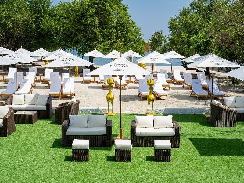 Escena de playa con sombrillas blancas, tumbonas, sofás y césped verde.