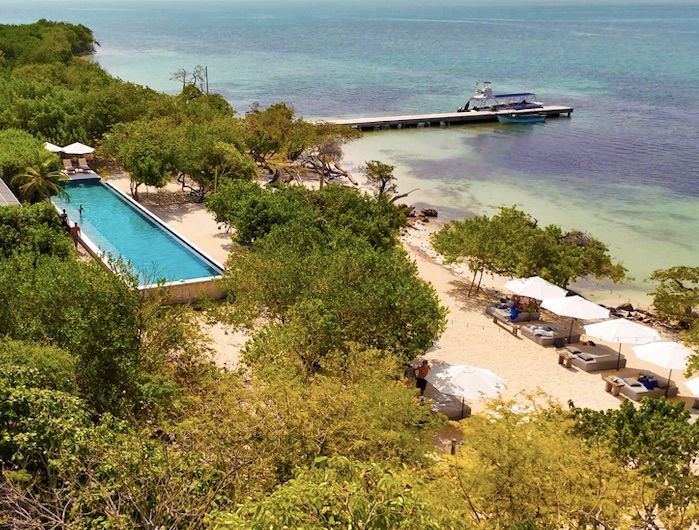 Complejo turístico frente a la playa con una larga piscina, muelle, arena blanca y agua turquesa.