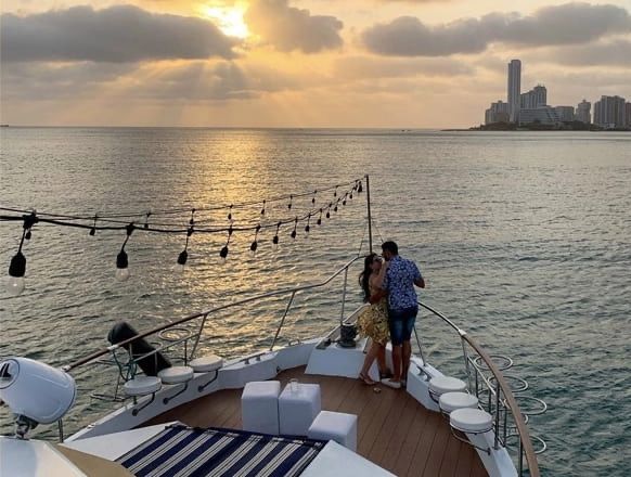 Pareja en la proa de un yate al atardecer, con el horizonte de la ciudad al fondo.