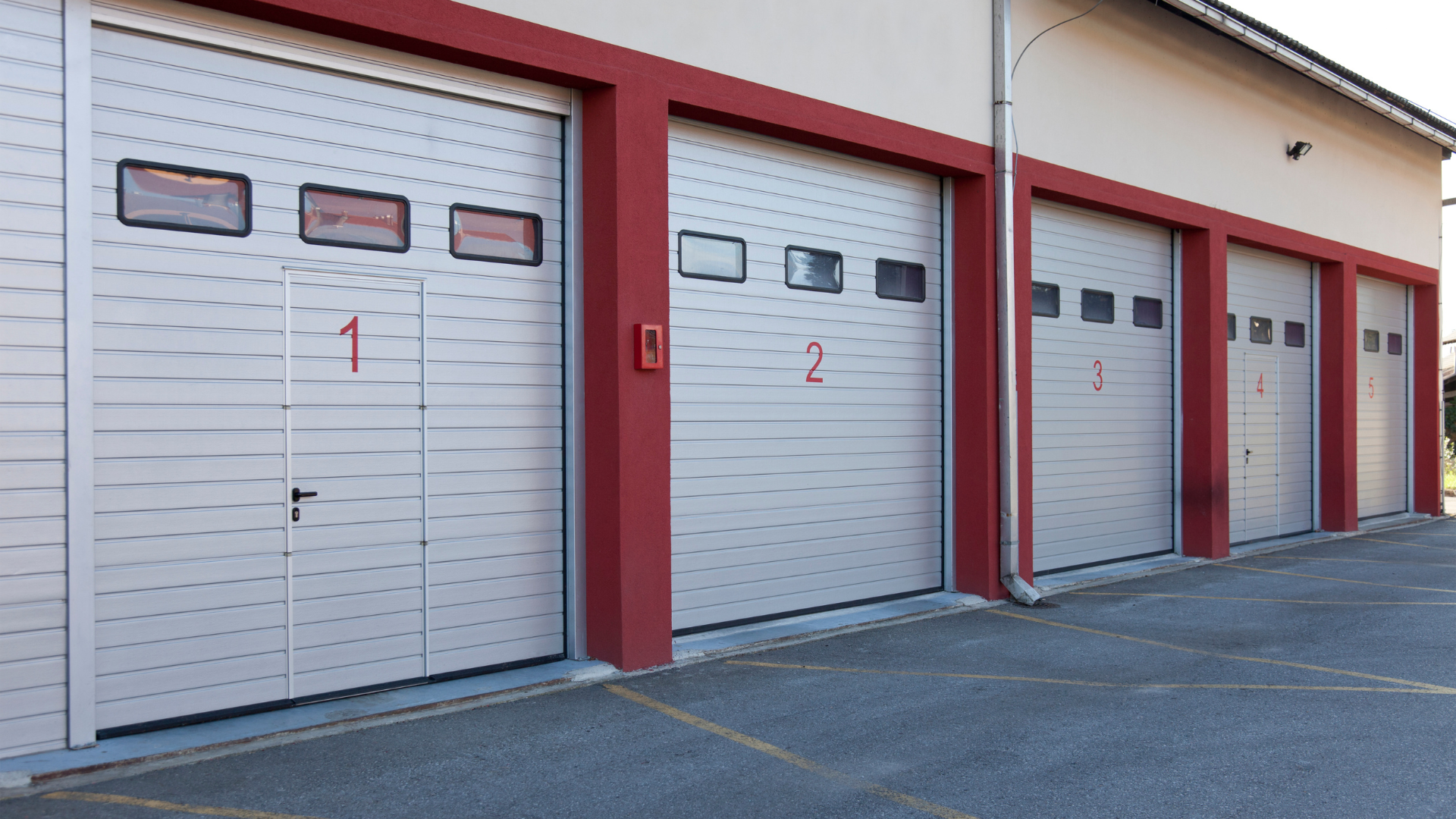 Four garage bays with light gray, paneled doors, red frames, and numbered 1-4, attached to a light-colored building.