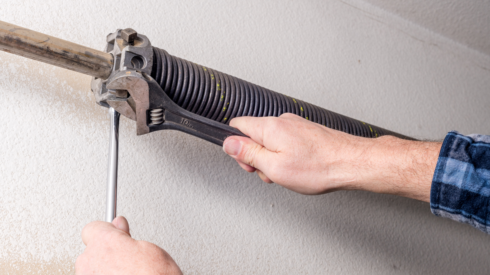 A person uses a wrench and a metal bar to adjust a garage door torsion spring mounted on a shaft.