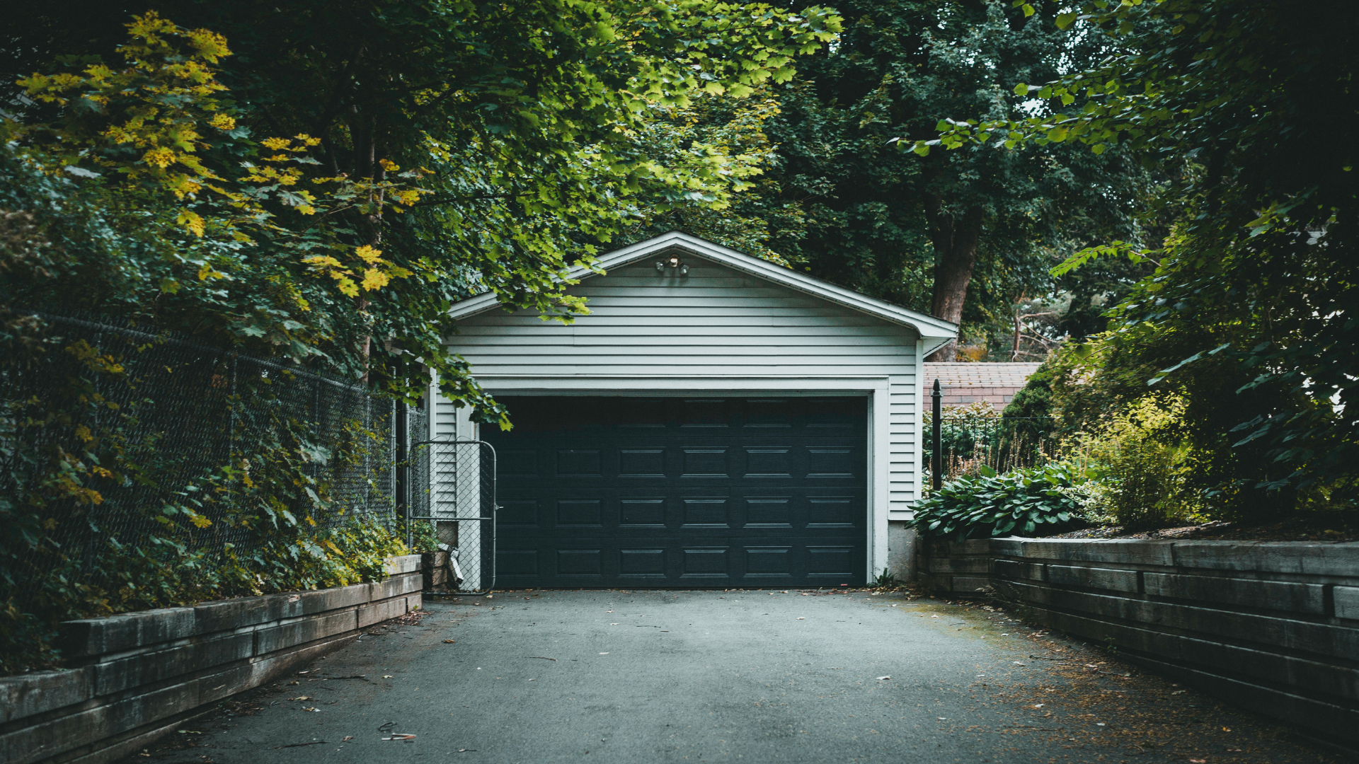 A white garage with a dark door at the end of a driveway, flanked by lush green trees and low stone walls.