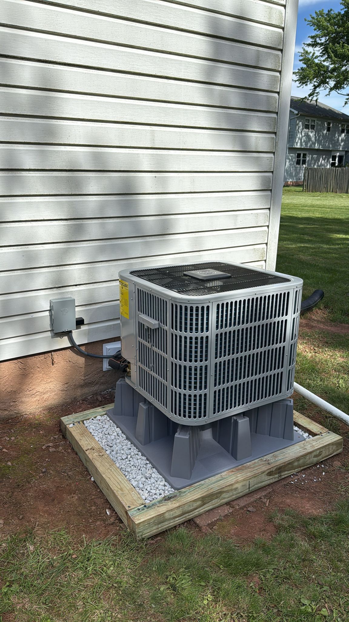 Outdoor air conditioning unit next to a light-colored building. Unit sits on a wooden platform with gravel.