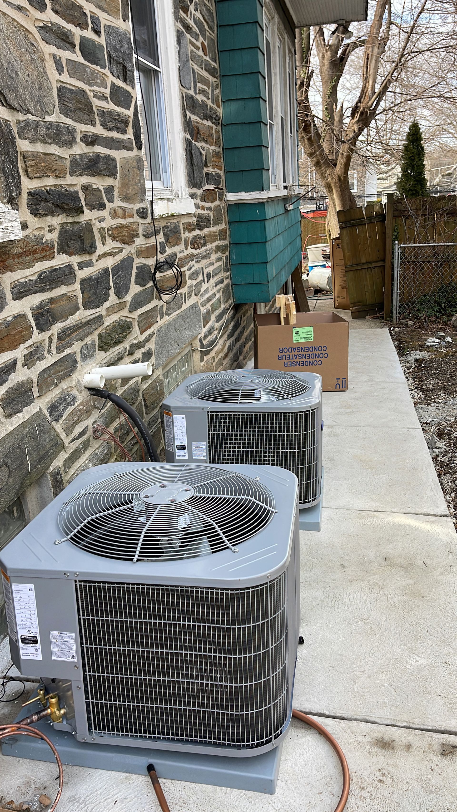 Two outdoor air conditioning units next to a stone wall and sidewalk.