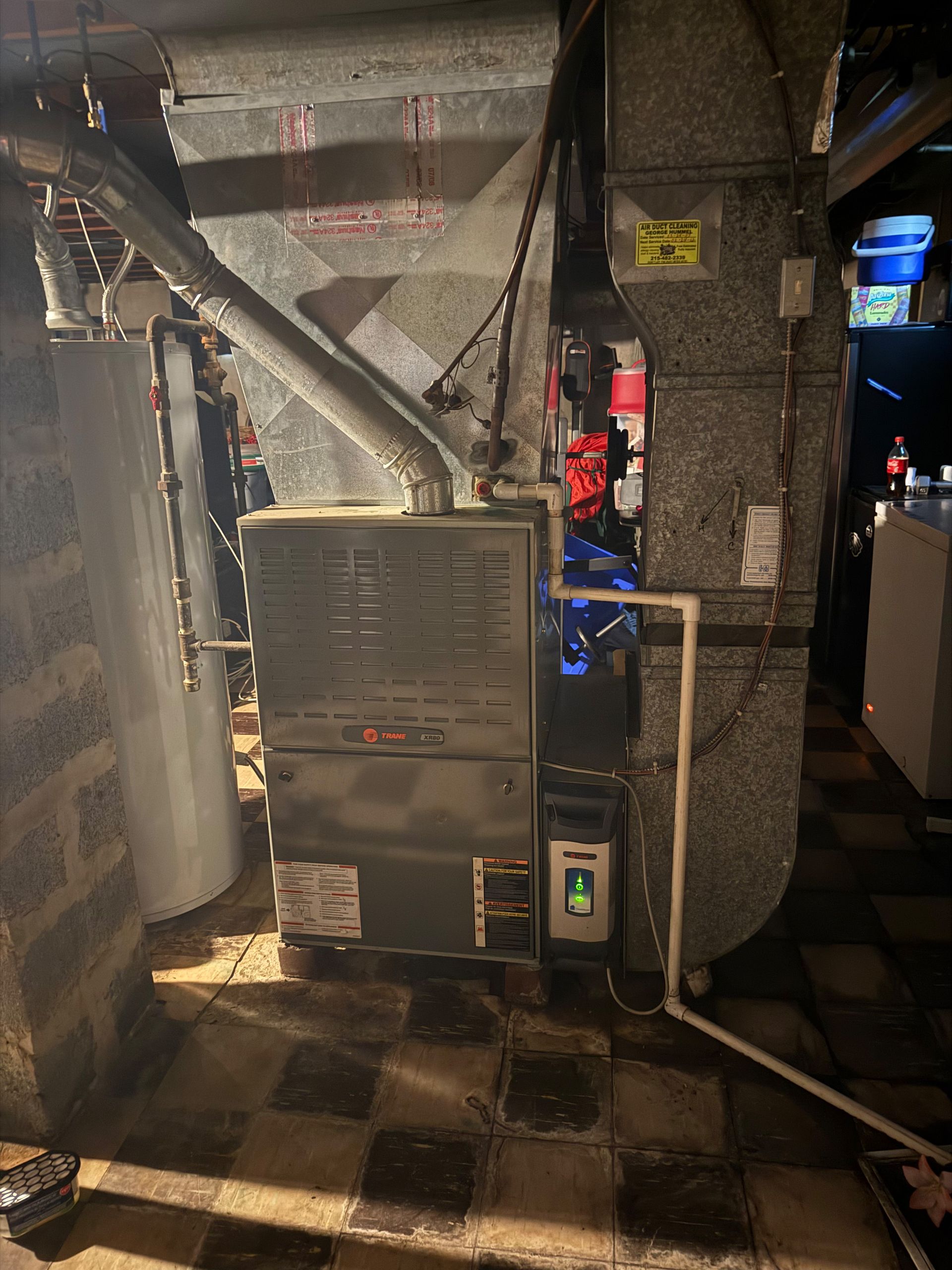 Furnace in a basement; gray metal unit with ductwork, water heater on the left, and a tiled floor.
