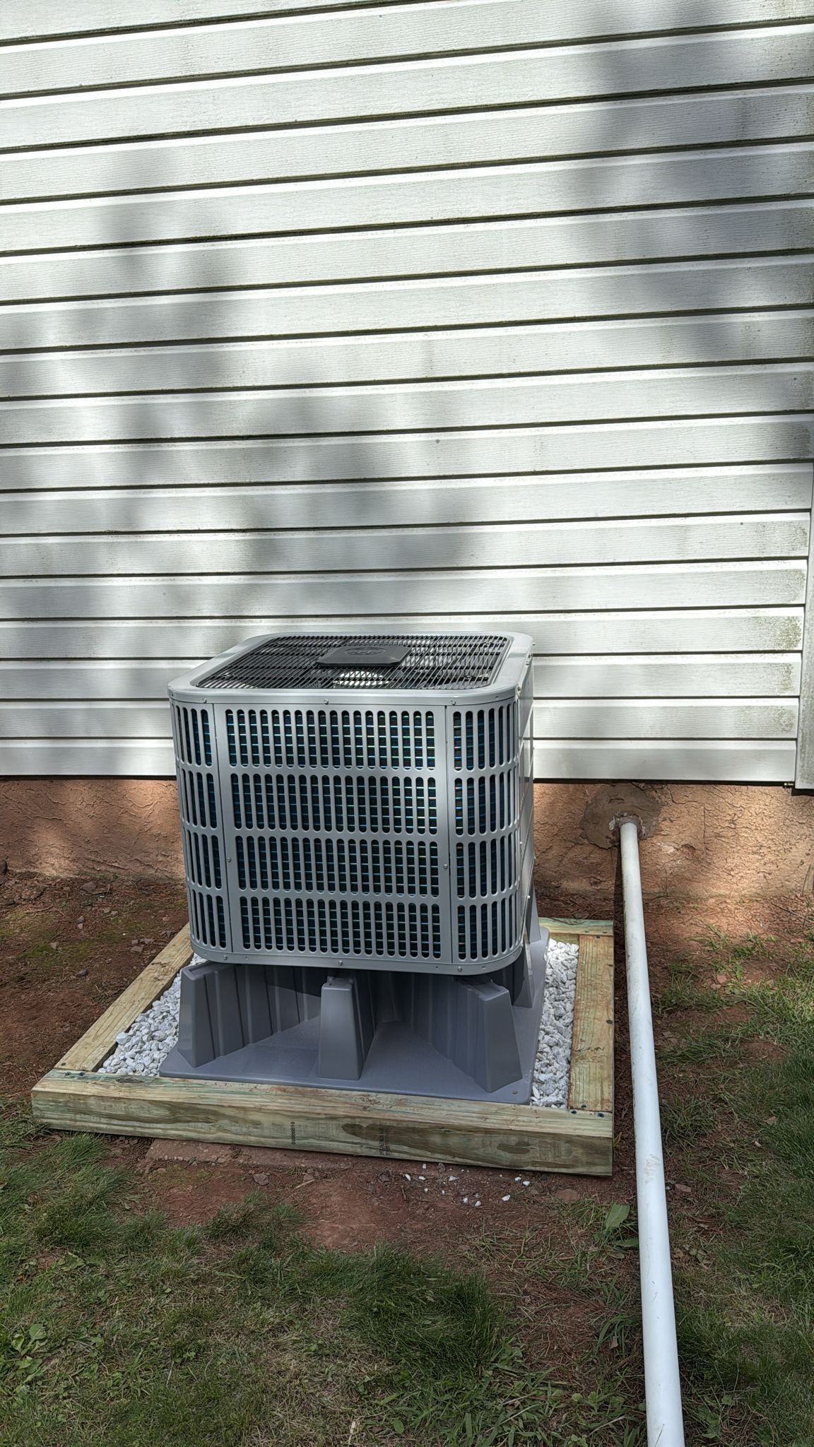 Air conditioning unit on a gravel bed next to a white house with horizontal siding; a white pipe is visible.