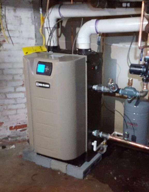 Tan and black boiler with blue digital display in a basement setting, connected to pipes.