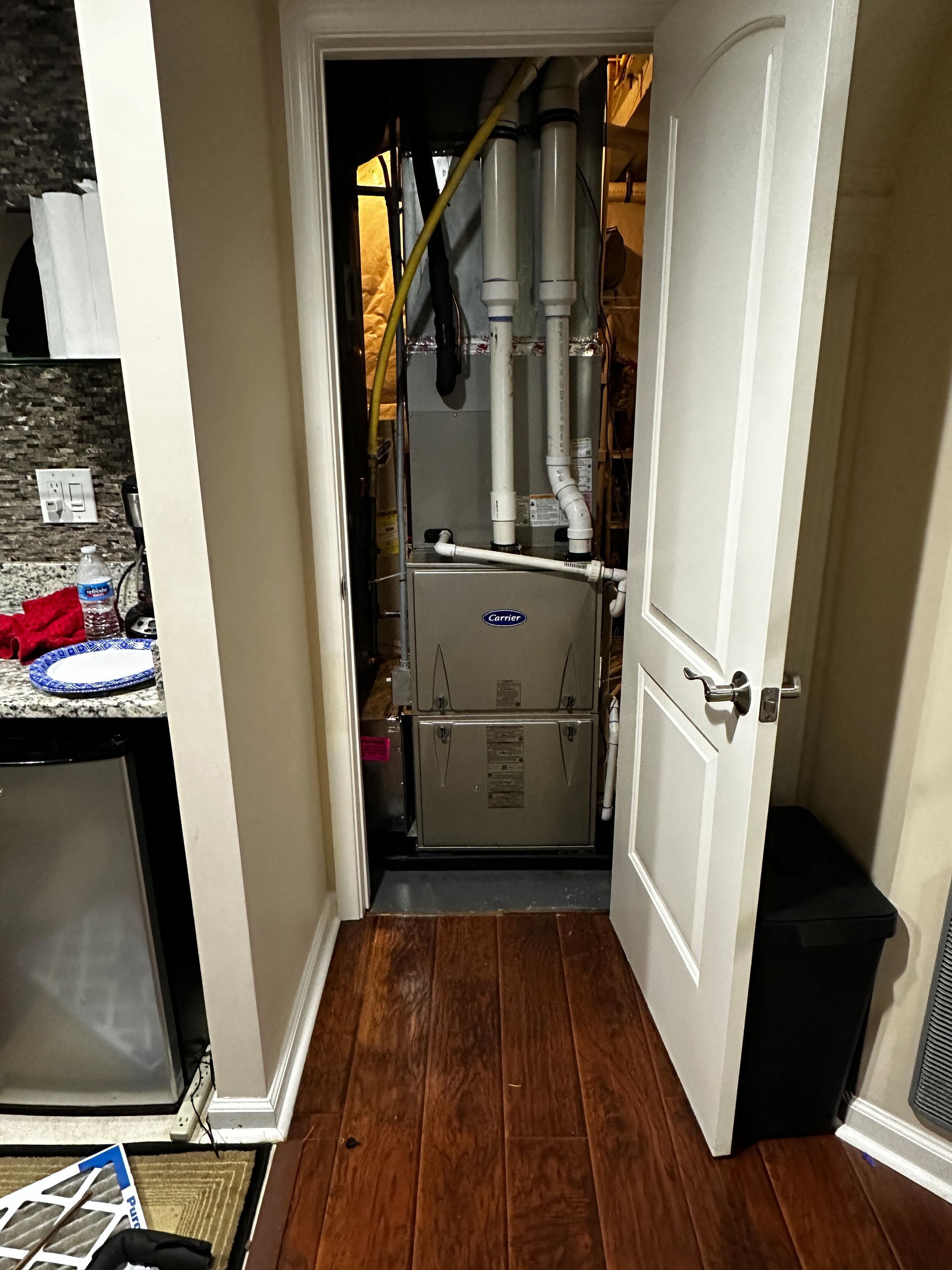 Furnace in a narrow closet with an open white door, dark wood floor, and black trash can.