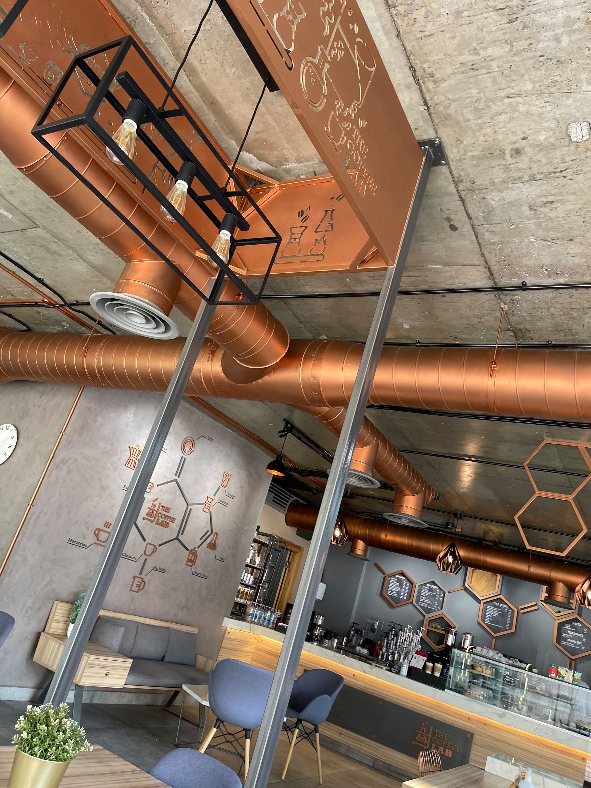 Cafe interior with copper pipes, exposed ceiling, geometric patterns, pendant lights, and bar area.