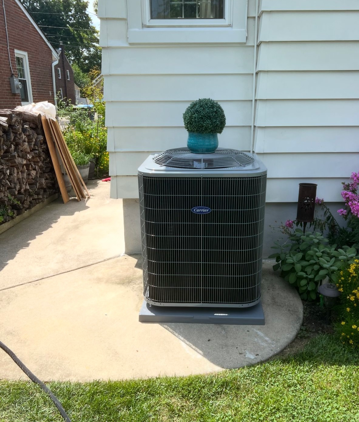 Air conditioning unit with a topiary on top, next to a white house with a curved concrete patio.