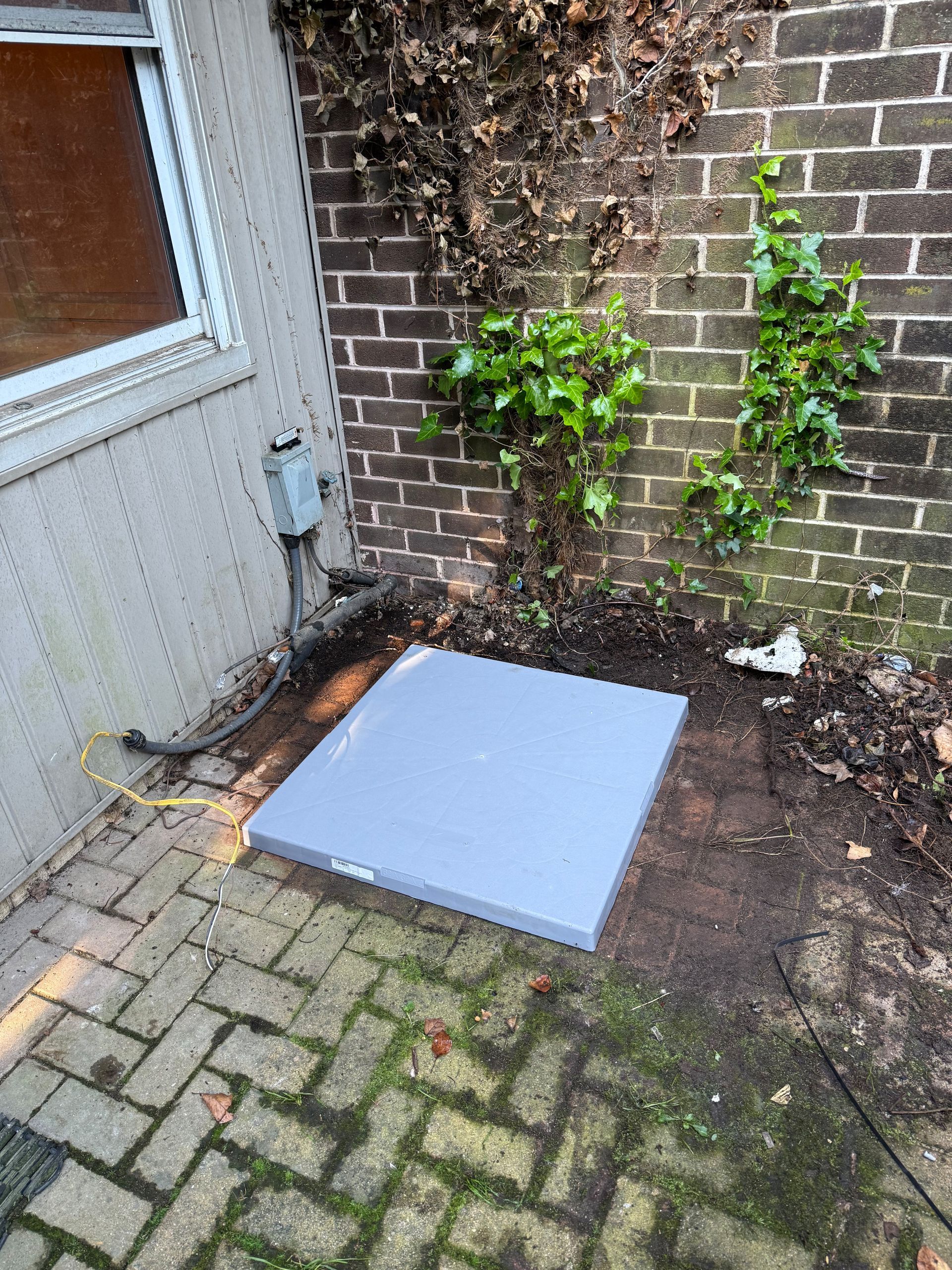 A gray square utility access cover sits on dirt next to a brick wall. A small electrical box is on a nearby wall.