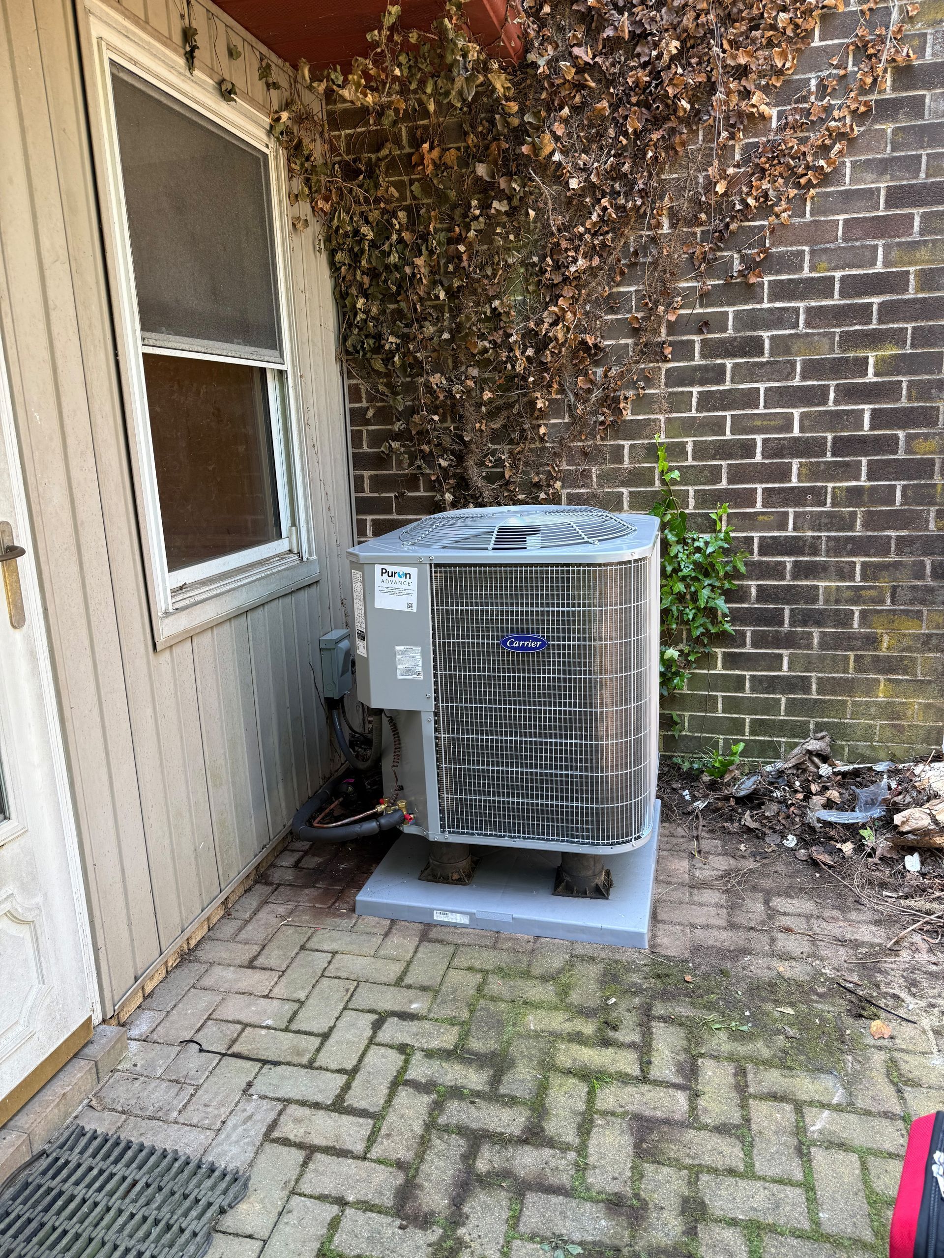 An air conditioning unit on a concrete pad next to a brick wall and a house.