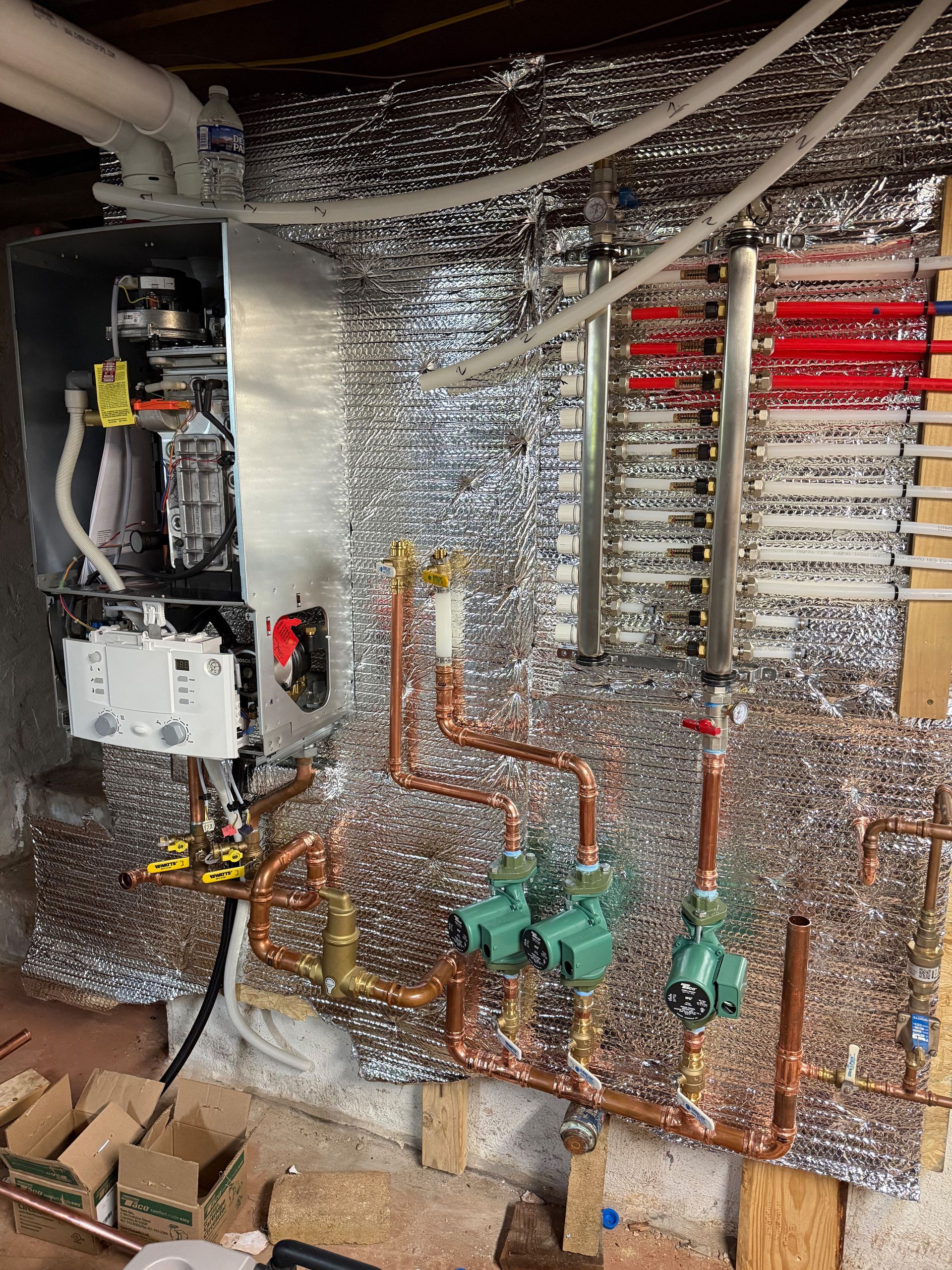 Hydronic heating system installation: boiler, copper pipes, pumps, and radiant heating manifolds on insulated wall.