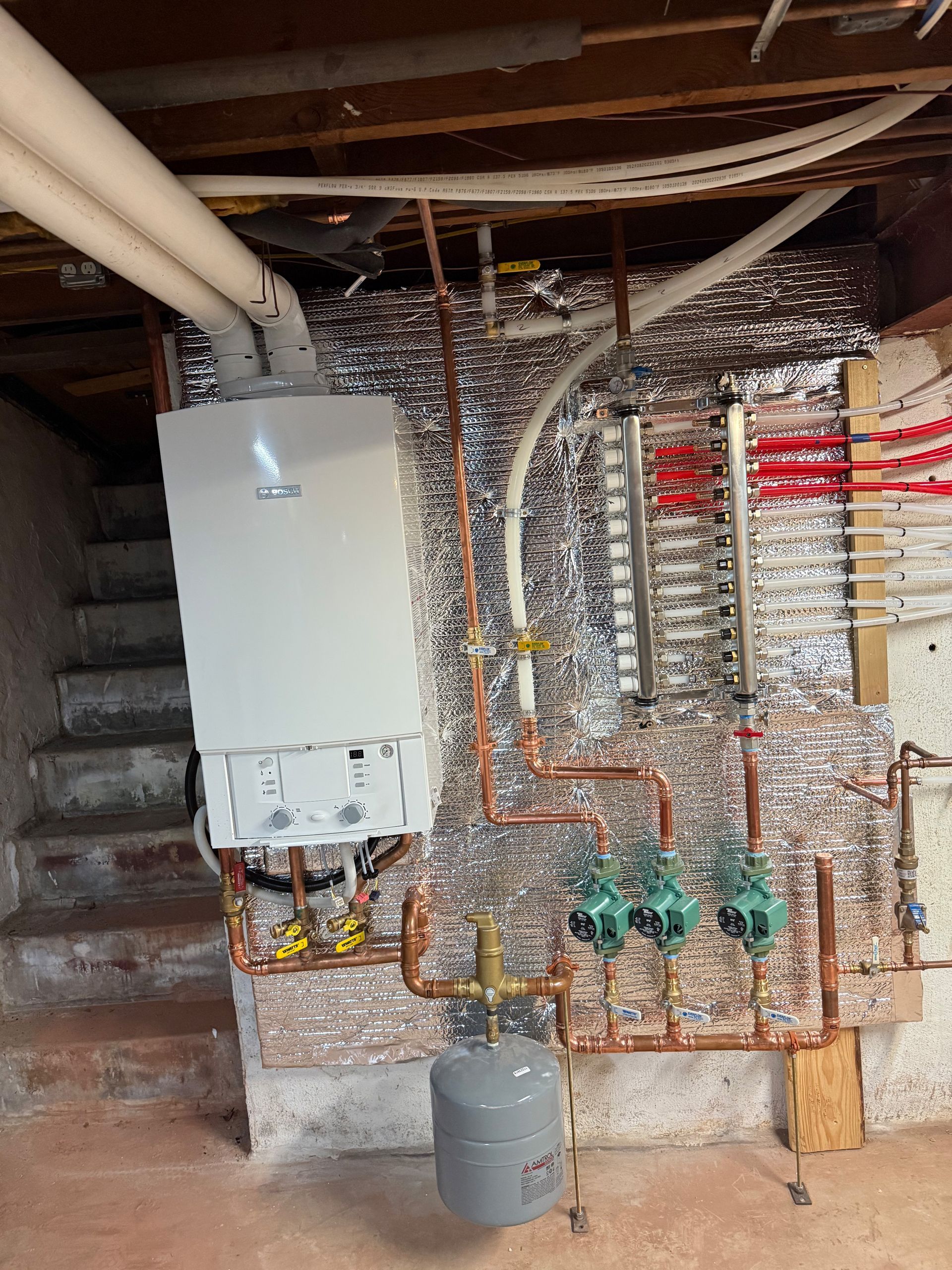 White wall-mounted boiler with copper pipes, circulation pumps, and expansion tank in a basement.