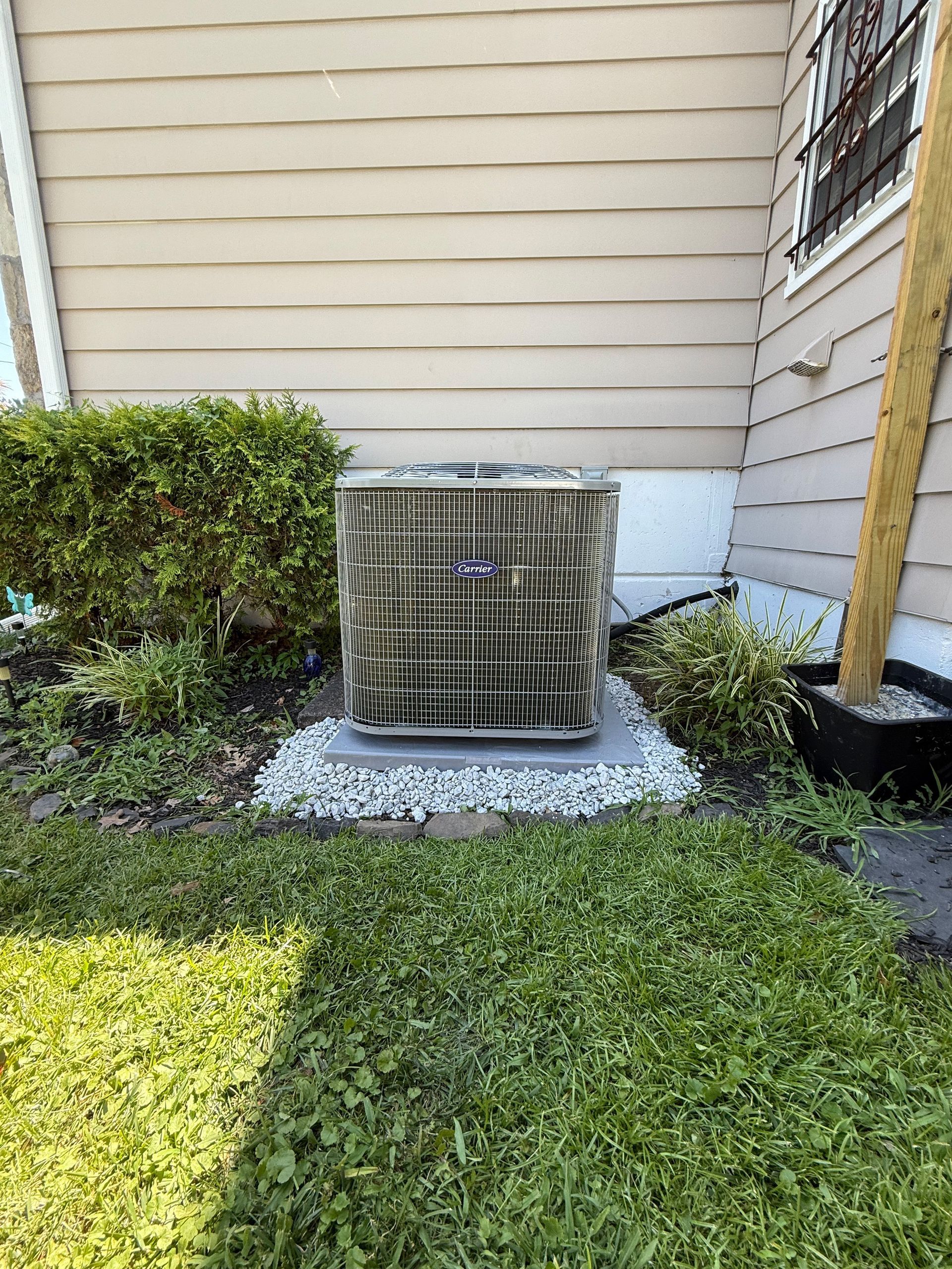AC unit on a gravel base next to a house with a bush and grass in a yard.