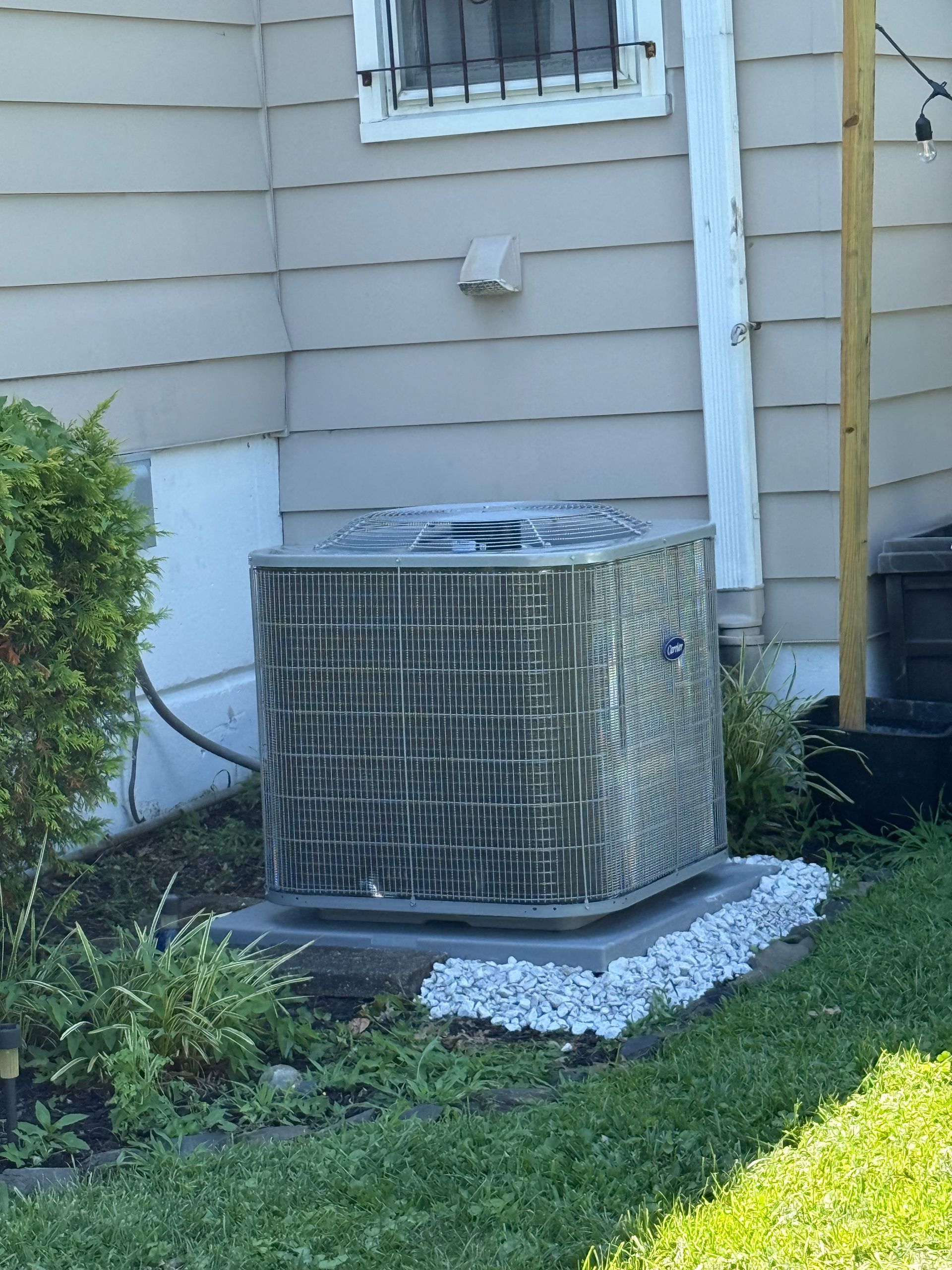 An outdoor air conditioning unit next to a light-colored building with a window, surrounded by small plants and gray gravel.