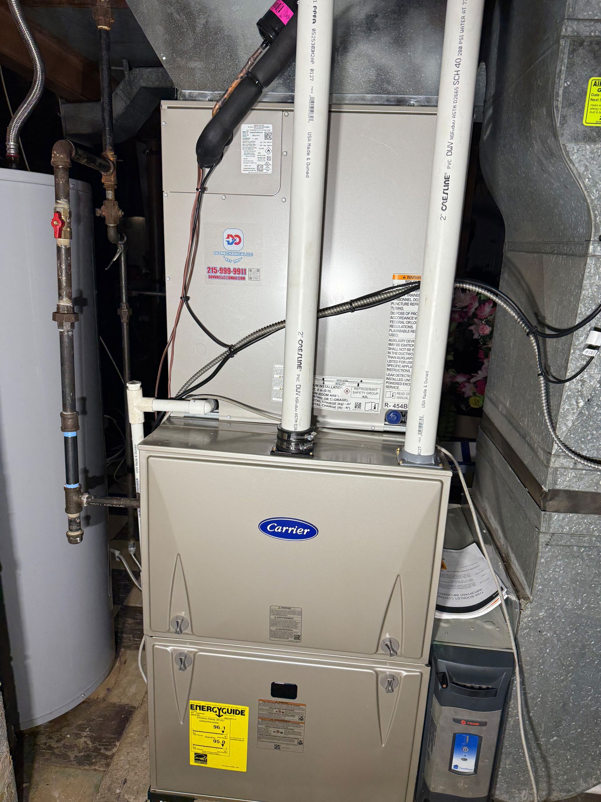 A furnace and air conditioning unit with white PVC pipes in a utility room next to a water heater.