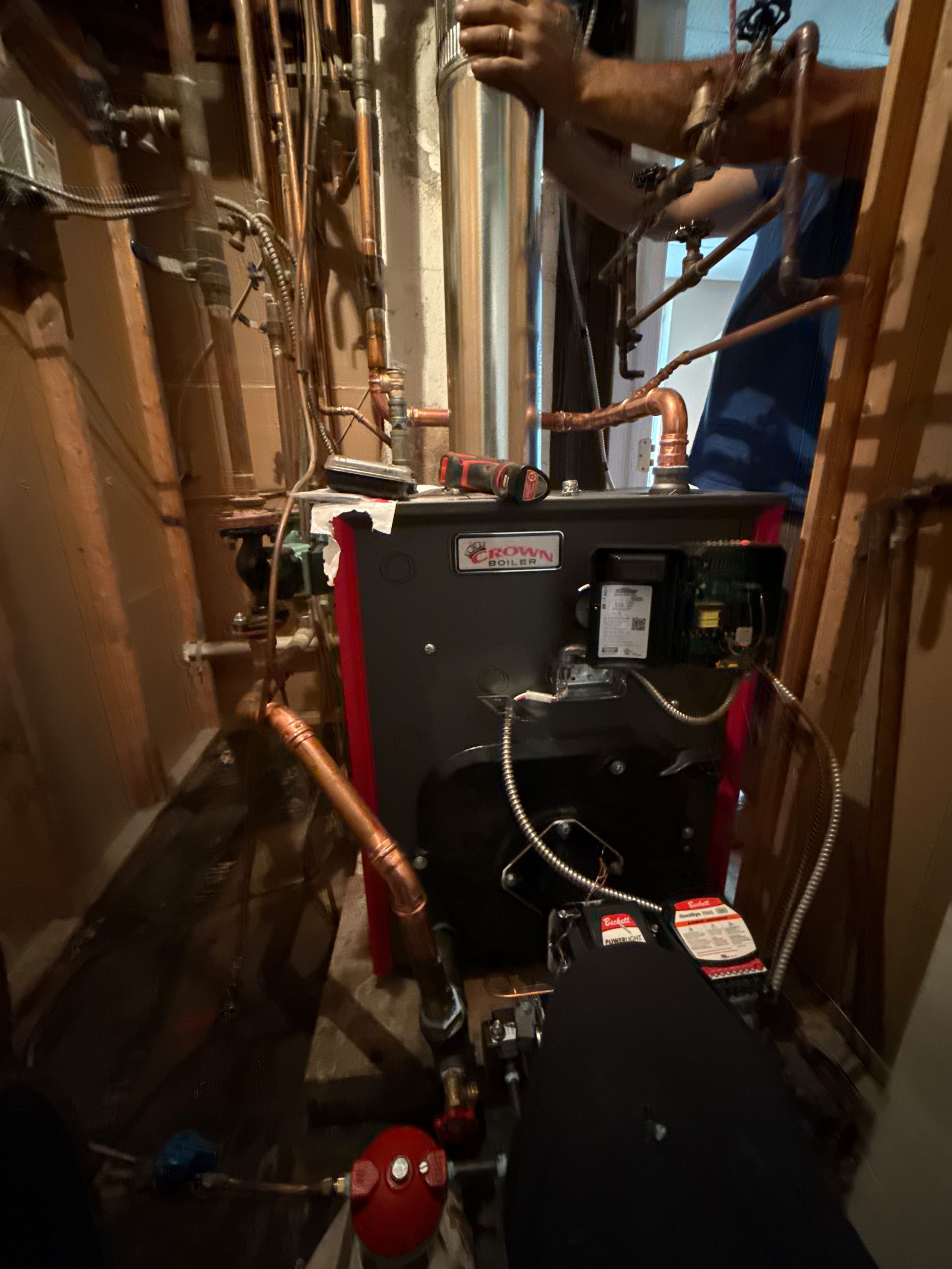 Plumber working on a dark gray boiler with copper pipes in a confined space.