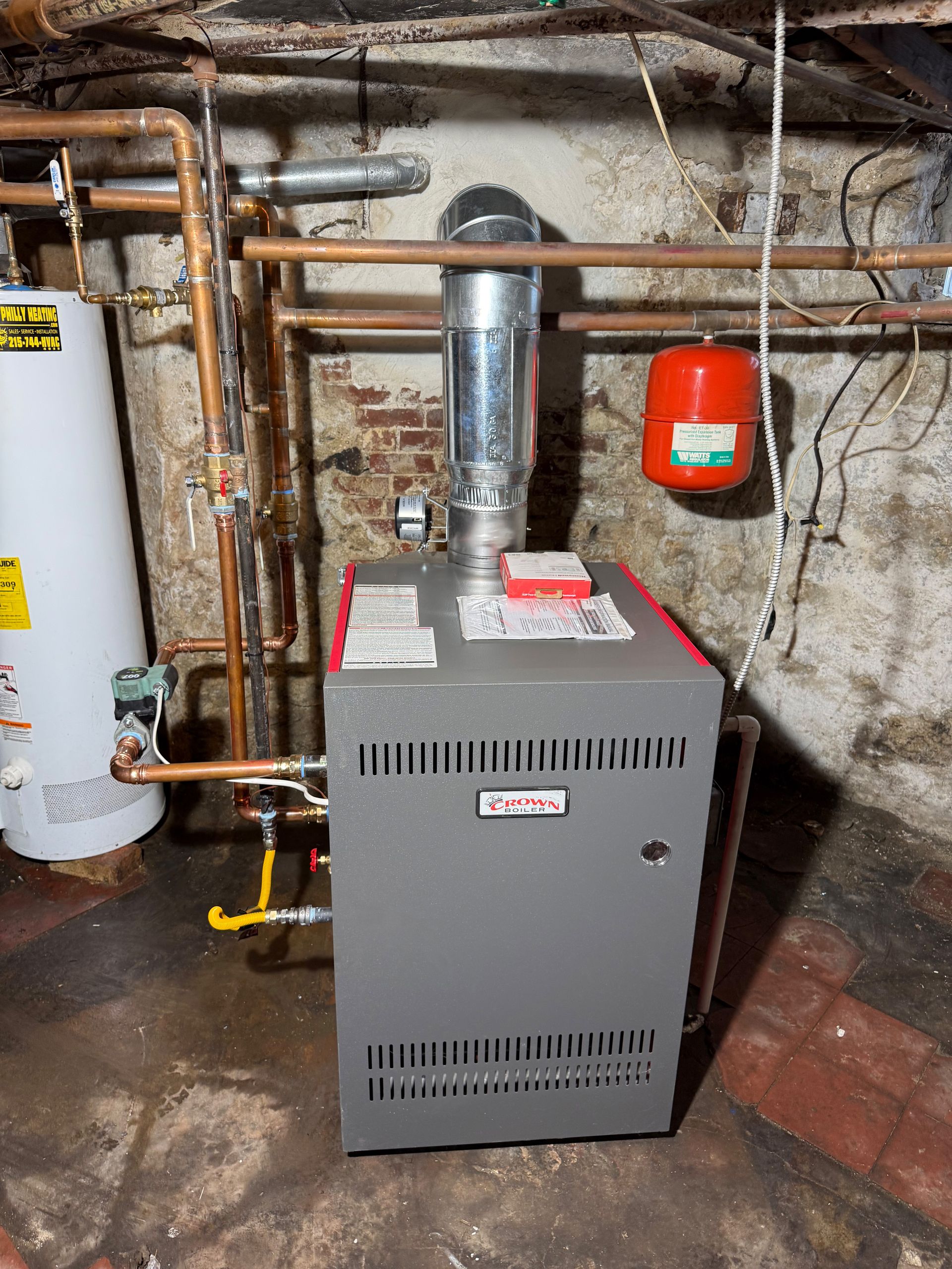 Furnace with silver vent pipe in basement, connected to copper pipes and a red expansion tank.