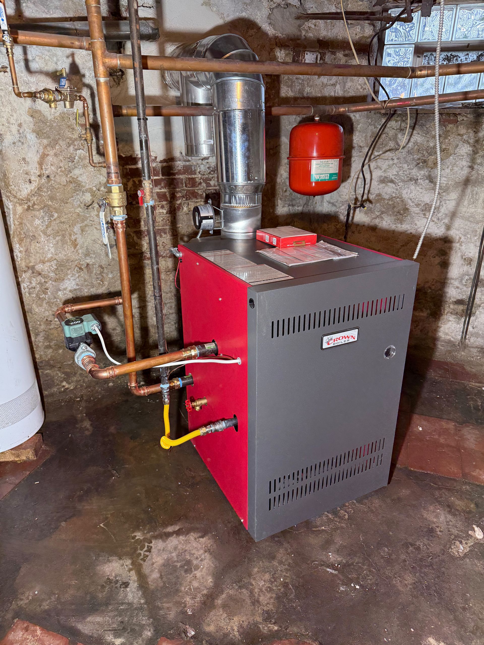 Red and gray boiler in a basement, connected to copper pipes, with a red expansion tank.