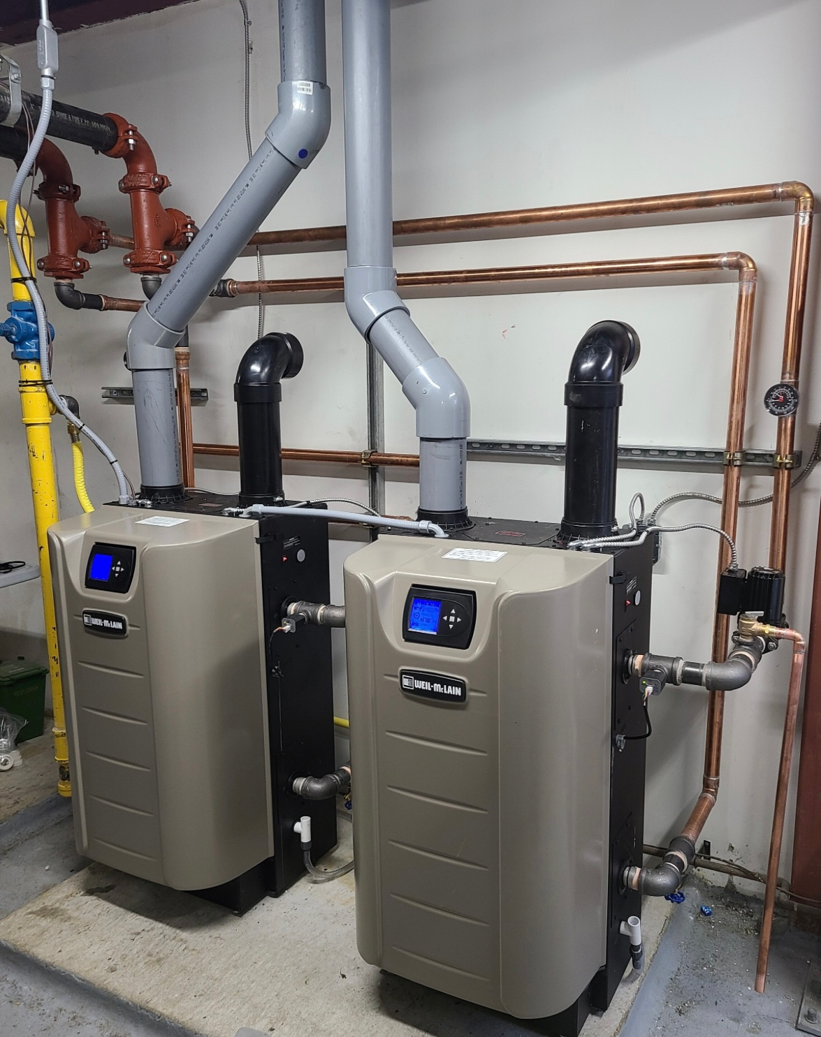 Two tan wall-mounted boilers with digital displays, connected by pipes, in a utility room.