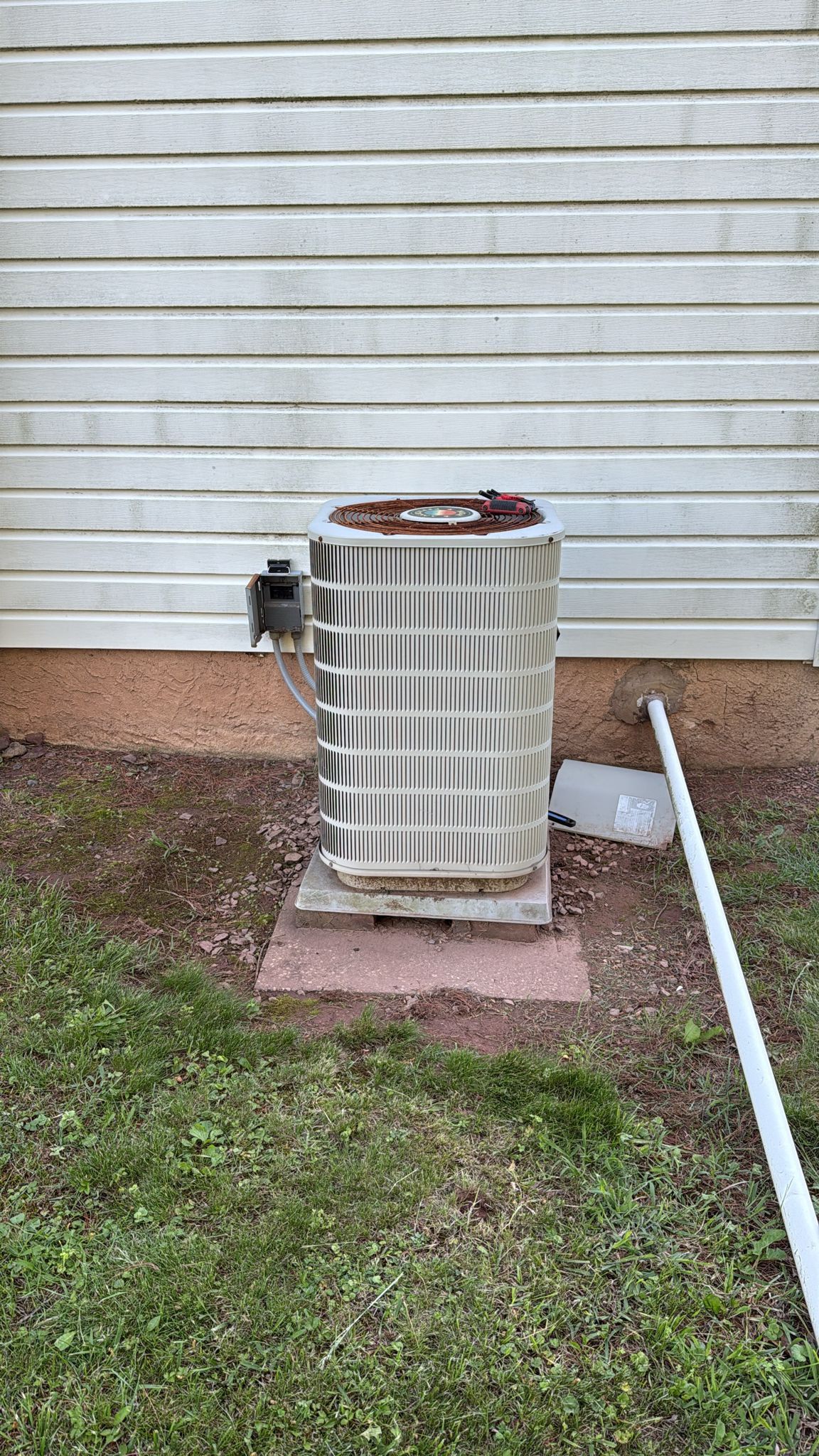 An air conditioner unit outside against a white-paneled building with a grassy area and red gravel.